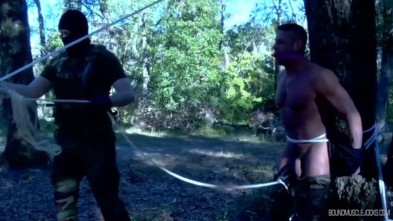 2 muscular military men are tied up to the trees outside
